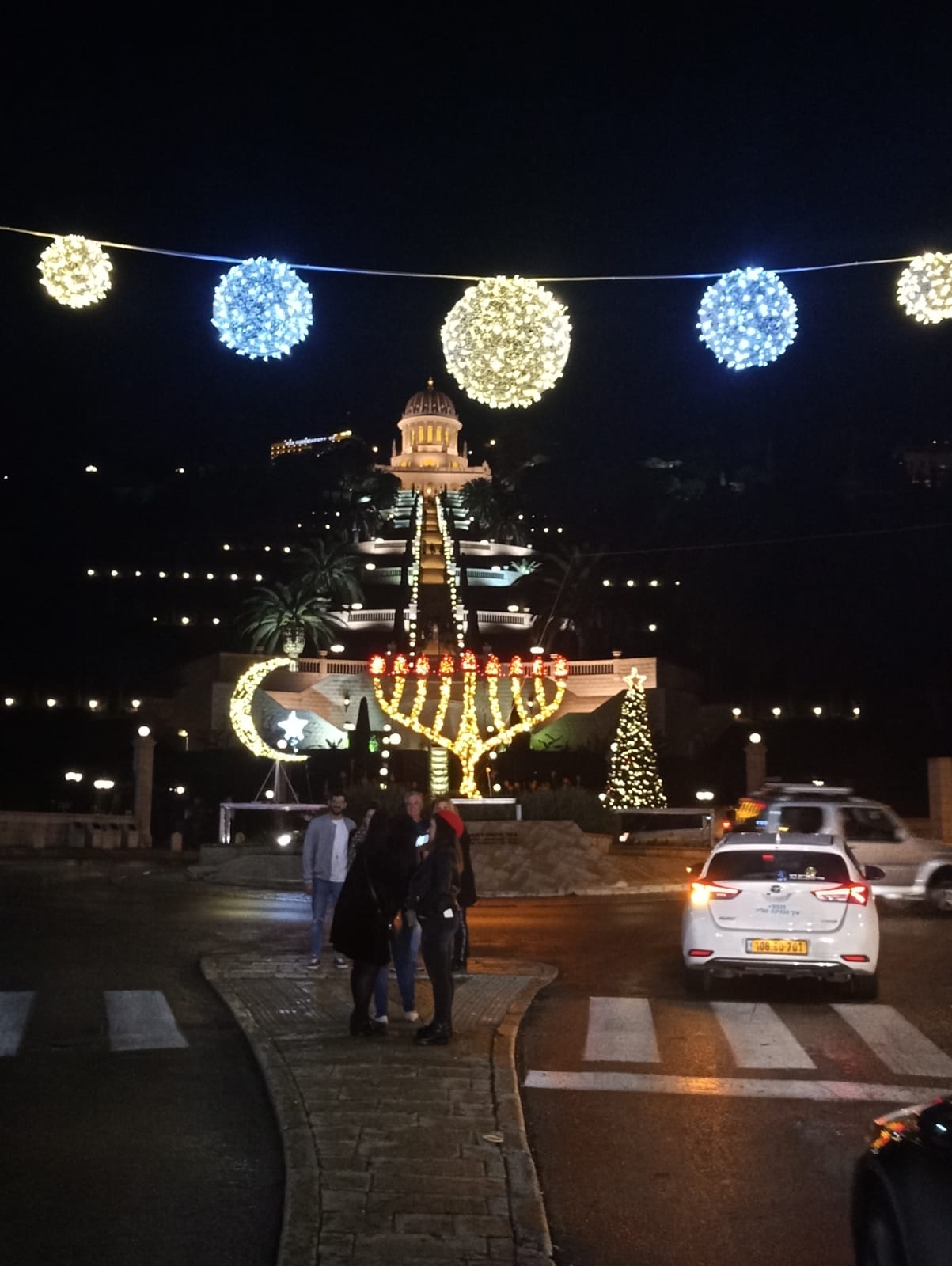 חנוכייה, סהר ועץ אשוח מוארים בלילה מול הגנים הבהאיים